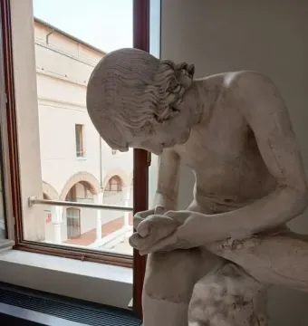 Boy with Thorn sculpture seen in profile, a light-colored boy leaning forward with his hands around his raised foot near a window.