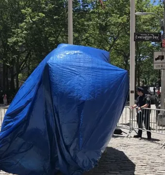 Charging Bull covered by a blue tarp on cobblestone ground, with police barriers and a street sign in the background.
