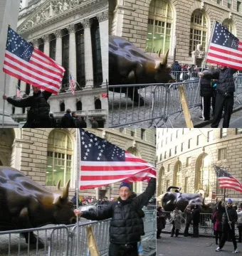 Charging Bull sculpture on a city street behind metal barricades, with people nearby holding American flags and classical buildings in the background.
