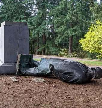 Harvey W. Scott statue toppled on its side beside a stone pedestal in a wooded area.