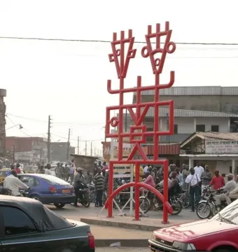 Njé Mo Yé, a large red geometric metal sculpture with two stylized heads, stands in a busy street setting.