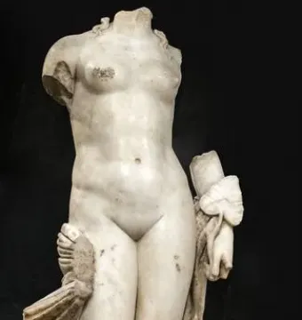 Headless white stone sculpture of Venus Unica on a metal base, with broken limbs and draped fabric against a dark background.