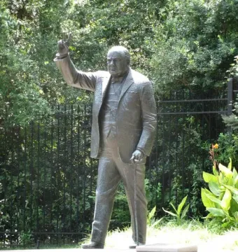 Winston Churchill bronze statue standing on a stone base and raising his right hand in a V for victory gesture while holding a cane.
