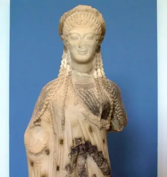 Chiotissa kore sculpture showing a female figure with braided hair and patterned draped garments on a red pedestal.