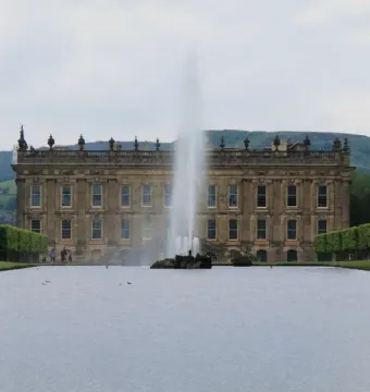 The Emperor Fountain shoots high in front of Chatsworth House, with green hills and trees under an overcast sky.