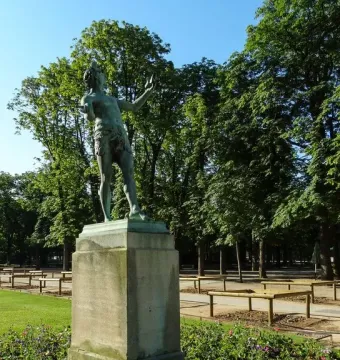 The Greek Actor sculpture on a tall stone pedestal in a park, with flowers and trees behind it.