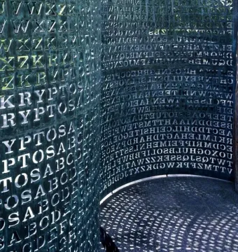 Kryptos, a curved metal sculpture covered with cut-out letters and numbers, with patterned shadows cast on the ground.