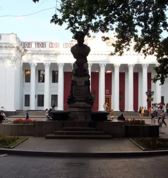 Pushkin bust on a tall pedestal in a paved plaza before a grand columned building.