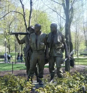 The Three Soldiers bronze sculpture standing in a park with trees, grass, and flowering bushes nearby.