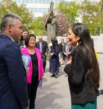 People gathered on a sunny plaza near the Japanese American Memorial to Patriotism During World War II, with a tall metallic sculpture and stone walls in the background.
