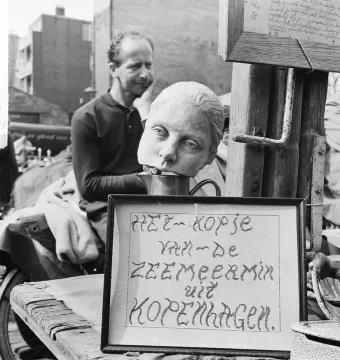 Detached head sculpture of The Little Mermaid resting in a cup, with a framed Dutch sign below it and a man seated in the background.