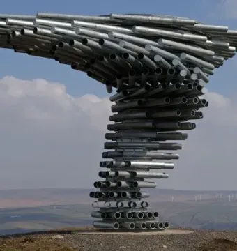 Singing Ringing Tree sculpture made of stacked silver metal pipes on a hillside with rolling landscape and distant wind turbines.
