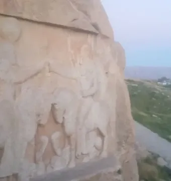 Ardashir Investiture Relief carved into a cliff, with horsemen and smaller figures overlooking green hills and a pathway.