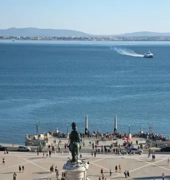 The Equestrian Statue of Joseph I on a tall stone pedestal in a sunny public square in Lisbon, surrounded by a fence and pedestrians.