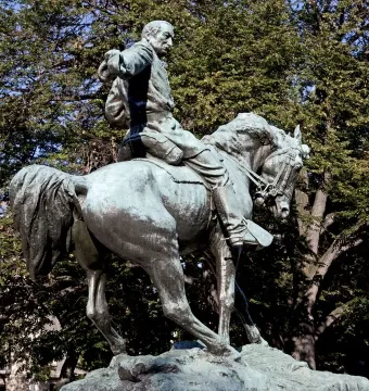 General Sheridan equestrian statue in bronze with a green patina, rearing on a horse outdoors among trees and blue sky.