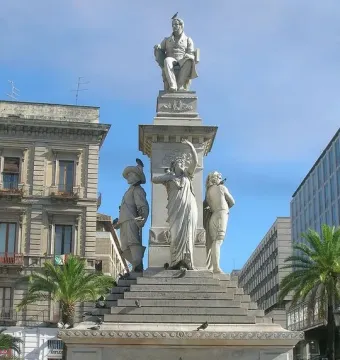 The Monument to Vincenzo Bellini, a tall white marble sculpture with a seated figure at the top and three standing allegorical figures below, surrounded by a decorative fence in a city square.
