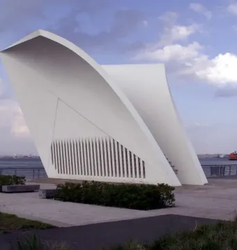 Postcards 9/11 Memorial with two white curved structures on a waterfront promenade, with a ferry and city skyline in the background.