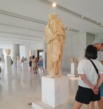 Weathered stone sculpture of Procne and Itys displayed on a pedestal in a museum gallery with visitors nearby.