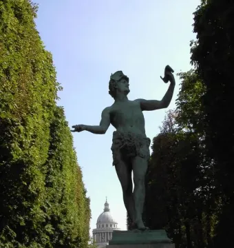 The Greek Actor statue on a stone pedestal with one arm raised holding a mask, set along a tree-lined path with a domed building in the distance.