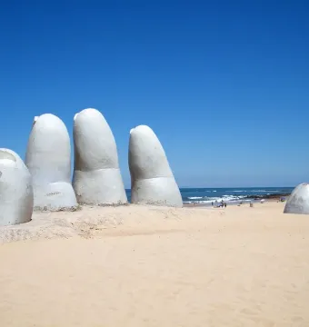 The Hand sculpture rising from the sand on a beach with the ocean and blue sky in the background.