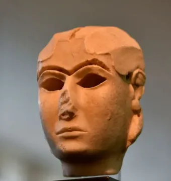 The Warka Mask, a light-toned ancient stone head with stylized facial features and hollow eye sockets on a dark pedestal.