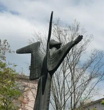 The Angel of Peace bronze statue with abstract wings and flowing robes, shown outdoors against bare trees and a cloudy sky.