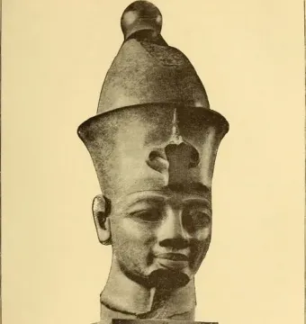 Sepia photograph of the Colossal Head of Amenhotep III, shown on a tiered pedestal with a tall crown and uraeus cobra emblem.