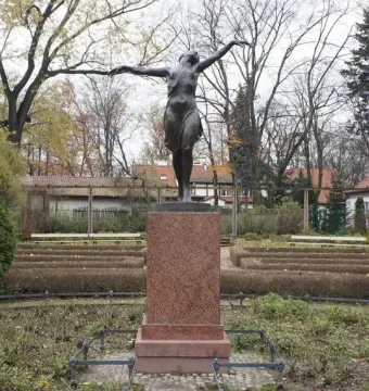 The Dancing Girl statue on a stone pedestal in a park with bushes and bare trees nearby.
