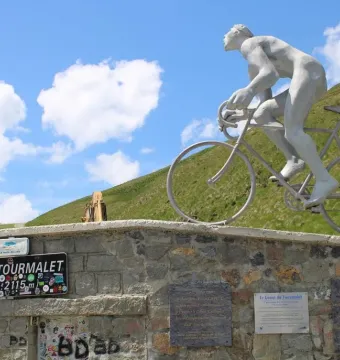 The Géant du Tourmalet statue of a cyclist on a bicycle beside a stone wall with mountain slopes and a blue sky behind it.