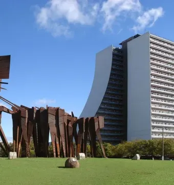 Monument to the Azoreans on a green lawn with a tall white building behind it under a blue sky.