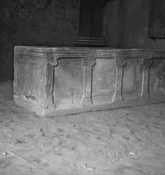 The Sarcophagus of Louis the German, a carved stone casket with fluted pilasters and spiral capitals, shown indoors on a rough floor.