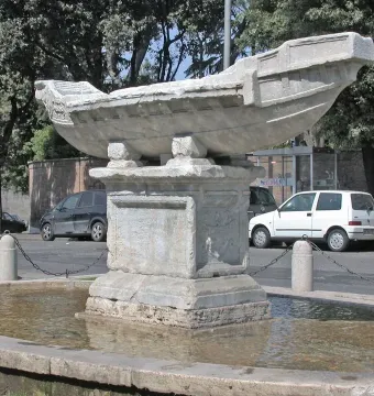 Fontana della Navicella stone boat fountain on a pedestal in a shallow basin, with trees and parked cars in the background.