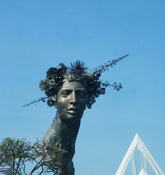 Reminiscencia sculpture showing a dark metal woman\'s head with an elaborate crown of flowers and branches against a blue sky.