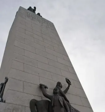 This is the Place Monument with bronze figures at its base and a tall stone shaft rising into the cloudy sky.