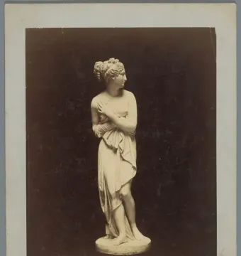 Sepia photograph of the Venus Italica marble statue, shown full-length against a dark background.