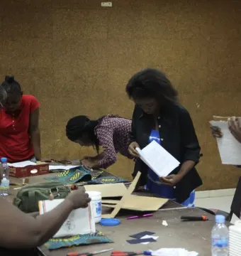 Women gathered around a table at a cartonería workshop, working with cardboard, fabric, scissors, and glue.