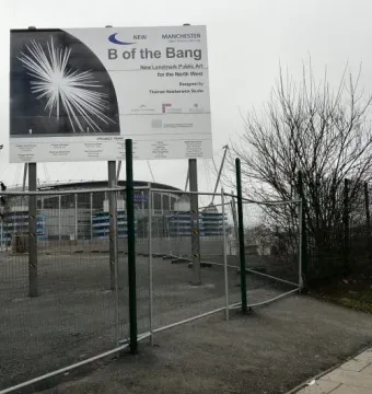 Information board for B of the Bang beside fenced gravel ground and a stadium building.