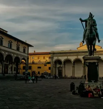Ferdinando I de\' Medici equestrian statue on a tall pedestal in a stone-paved plaza with historic arched porticos and scattered people nearby.