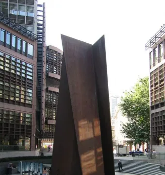 Fulcrum sculpture in a paved plaza with people walking nearby and office buildings in the background.
