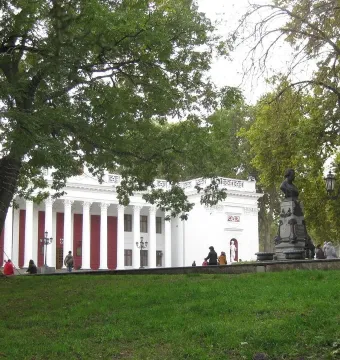 Pushkin bust on an ornate pedestal in a park with trees, a lamppost, and a columned building behind it.