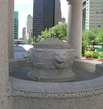 Bagley Memorial Fountain with carved lion heads around the central basin, framed by stone columns and a city skyline in the background.