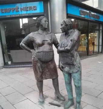 Monument to the Unknown Woman Worker with two bronze women standing on a paved street outside a Caffè Nero.