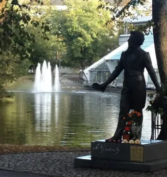 Pushkin statue on a stone pedestal near a pond with fountains, with trees and a modern building in the background.