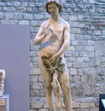 Adam statue holding leaves over his groin beside a stone wall and tree trunk.