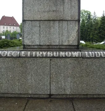 Lower section of the Mickiewicz Monument with a grey granite pedestal and carved inscription, surrounded by bushes and wet paving stones.