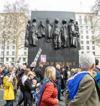 The Monument to the Women of World War II stands behind a crowd of protesters in a city square.