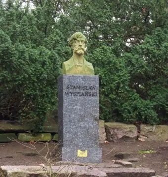Stanisław Wyspiański bust on a tall stone pedestal in a leafy outdoor setting.