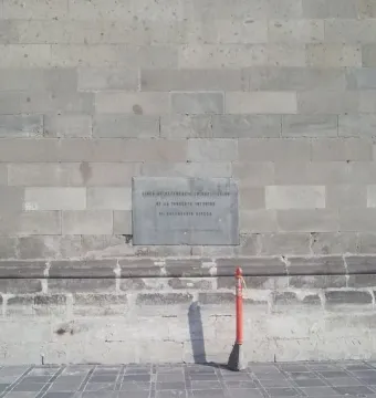 Spanish plaque set into a light stone wall marking the Aztec Sun Stone reference point, with an orange post and metal box nearby.