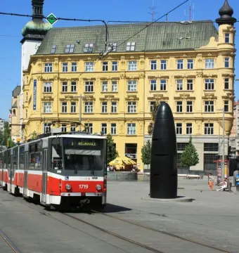 The Brno astronomical clock stands in a city square as a tram passes in front of it, with historic buildings in the background.