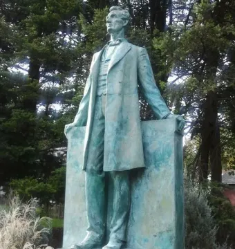 Lincoln the Lawyer bronze statue of Abraham Lincoln standing outdoors on a pedestal with evergreen trees behind it.